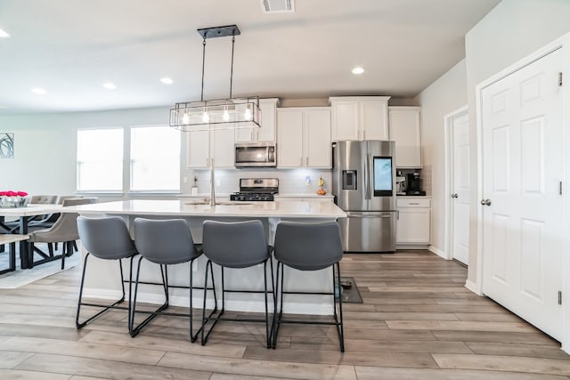 kitchen featuring white cabinets, an island with sink, pendant lighting, light wood-type flooring, and appliances with stainless steel finishes