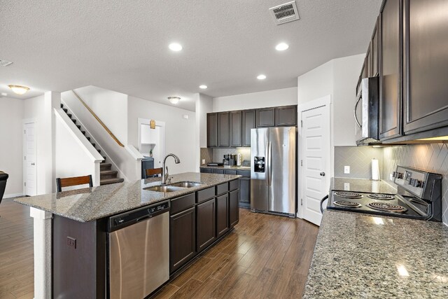 kitchen with stainless steel appliances, dark hardwood / wood-style flooring, light stone counters, and a center island with sink