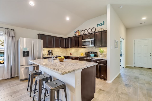 kitchen with lofted ceiling, backsplash, a kitchen island with sink, light stone countertops, and stainless steel appliances