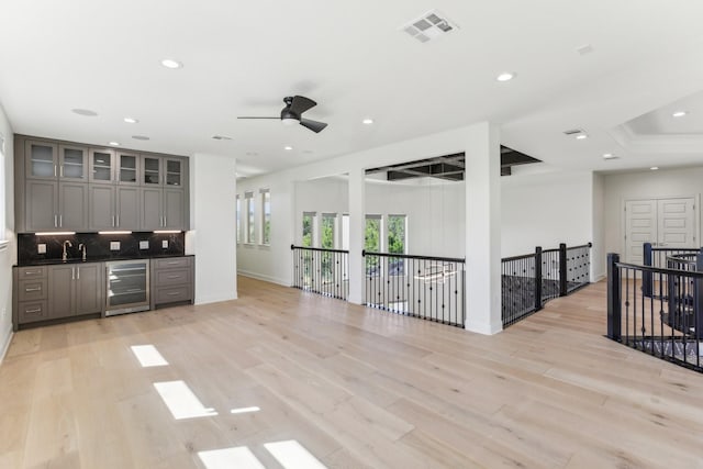 interior space with light wood-type flooring, gray cabinets, ceiling fan, beverage cooler, and backsplash