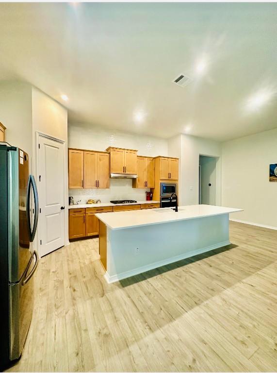 kitchen with sink, black fridge, a center island with sink, light wood-type flooring, and stainless steel microwave