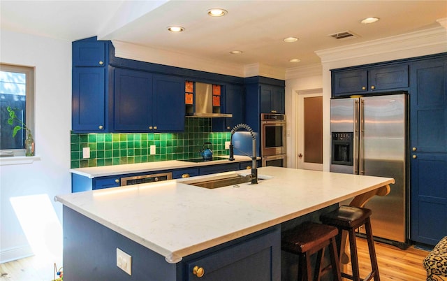 kitchen featuring blue cabinets, stainless steel appliances, wall chimney exhaust hood, and an island with sink