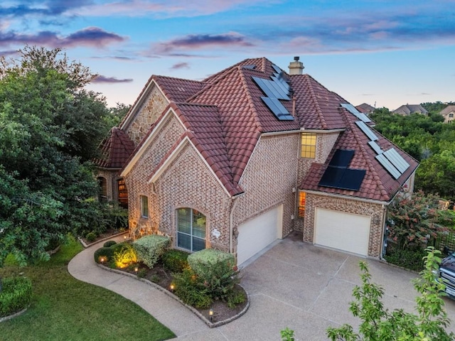 view of front facade with roof mounted solar panels, driveway, a tile roof, a chimney, and brick siding
