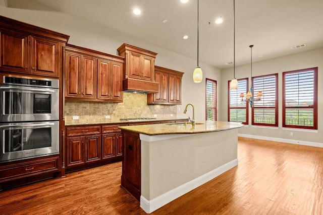 kitchen featuring appliances with stainless steel finishes, hanging light fixtures, sink, an island with sink, and light stone counters