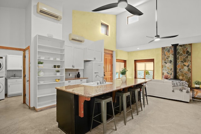 kitchen with white cabinets, light stone countertops, an AC wall unit, an island with sink, and stacked washer / dryer