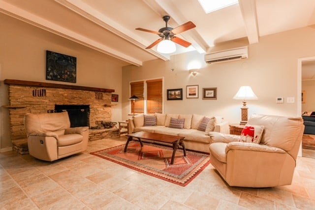 living room with light tile patterned floors, ceiling fan, beamed ceiling, a stone fireplace, and a wall unit AC