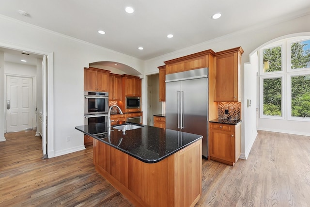 kitchen with an island with sink, built in appliances, crown molding, sink, and backsplash