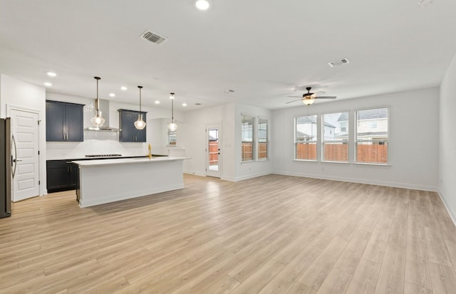 kitchen featuring wall chimney range hood, tasteful backsplash, an island with sink, hanging light fixtures, and light wood-type flooring