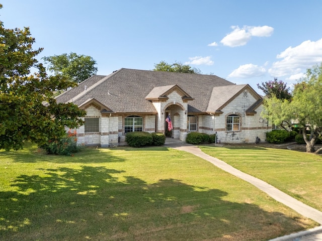 french country home with a shingled roof, a front yard, and stone siding