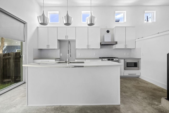 kitchen with decorative light fixtures, wall chimney range hood, appliances with stainless steel finishes, an island with sink, and white cabinets