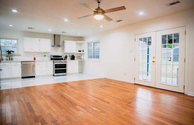 kitchen with french doors, white cabinetry, light wood-type flooring, appliances with stainless steel finishes, and wall chimney range hood