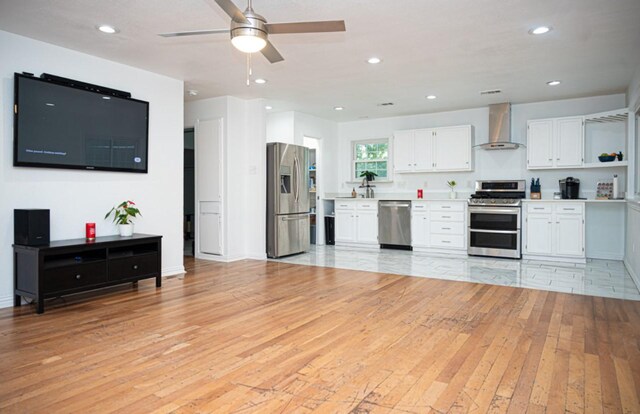 kitchen featuring wall chimney range hood, light wood-type flooring, ceiling fan, stainless steel appliances, and white cabinets