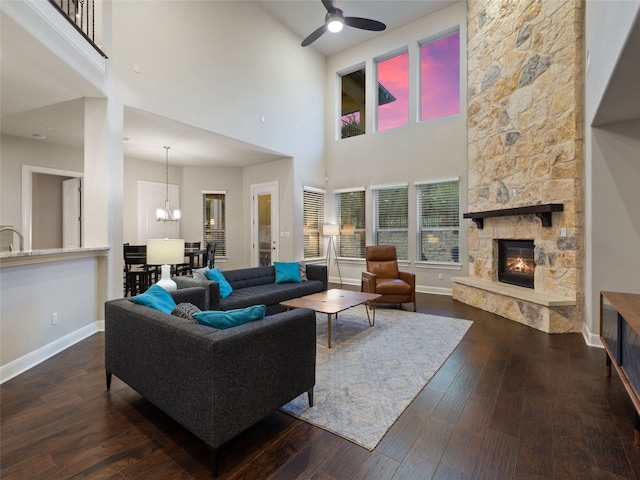 living area with dark wood-style flooring, a stone fireplace, baseboards, and ceiling fan with notable chandelier