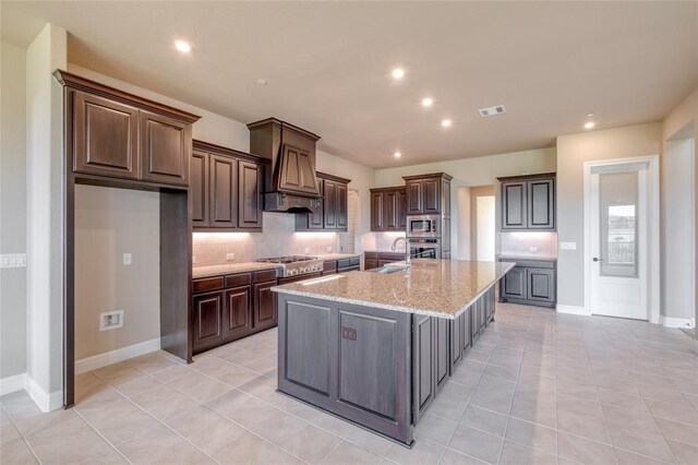 kitchen featuring custom exhaust hood, dark brown cabinets, stainless steel appliances, decorative backsplash, and a kitchen island with sink