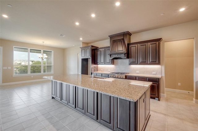 kitchen with backsplash, premium range hood, light stone countertops, sink, and a center island with sink