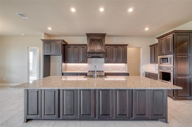 kitchen with appliances with stainless steel finishes, tasteful backsplash, light stone counters, custom exhaust hood, and an island with sink