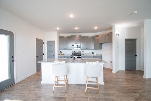 kitchen with a kitchen breakfast bar, light stone countertops, gray cabinets, stainless steel appliances, and a center island with sink