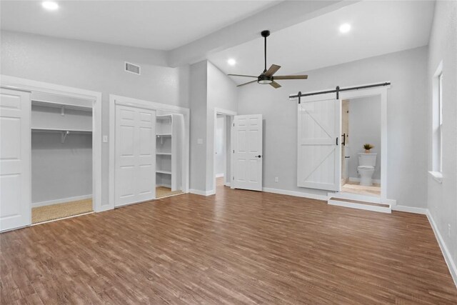 unfurnished bedroom with wood-type flooring, ceiling fan, high vaulted ceiling, a barn door, and ensuite bathroom