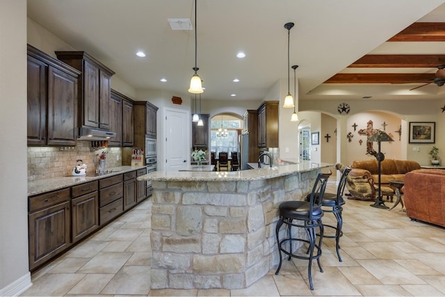 kitchen with light stone countertops, a kitchen breakfast bar, beamed ceiling, backsplash, and decorative light fixtures