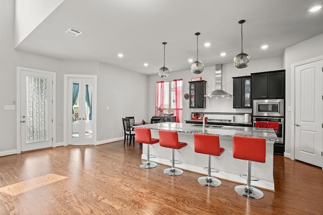 kitchen featuring wall chimney range hood, wood-type flooring, stainless steel appliances, and an island with sink