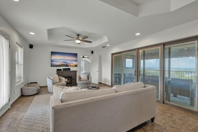 living area featuring baseboards, a raised ceiling, ceiling fan, a lit fireplace, and recessed lighting