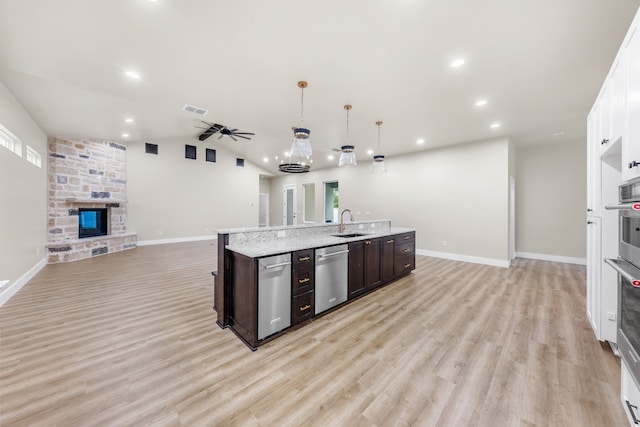 kitchen featuring a stone fireplace, dark brown cabinets, hanging light fixtures, stainless steel dishwasher, and a kitchen island with sink