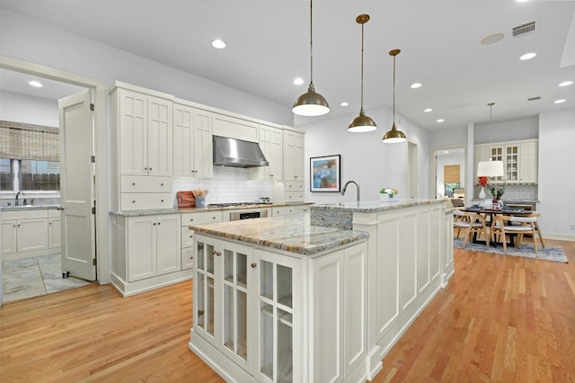 kitchen with white cabinets, an island with sink, hanging light fixtures, exhaust hood, and light hardwood / wood-style floors