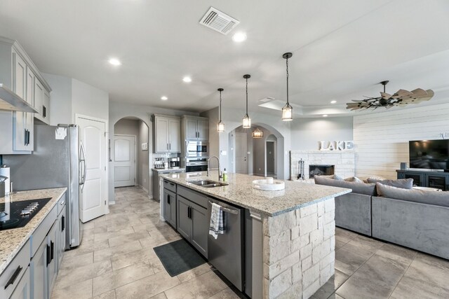 kitchen with gray cabinetry, a stone fireplace, an island with sink, and ceiling fan