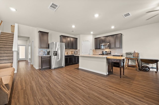 kitchen featuring dark wood-type flooring, dark brown cabinetry, appliances with stainless steel finishes, a kitchen island with sink, and backsplash