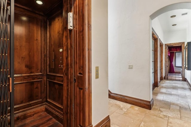 corridor featuring stone tile flooring, visible vents, baseboards, arched walkways, and elevator