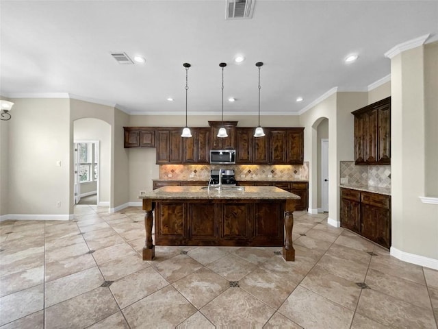 kitchen with appliances with stainless steel finishes, hanging light fixtures, light stone counters, dark brown cabinets, and a center island with sink