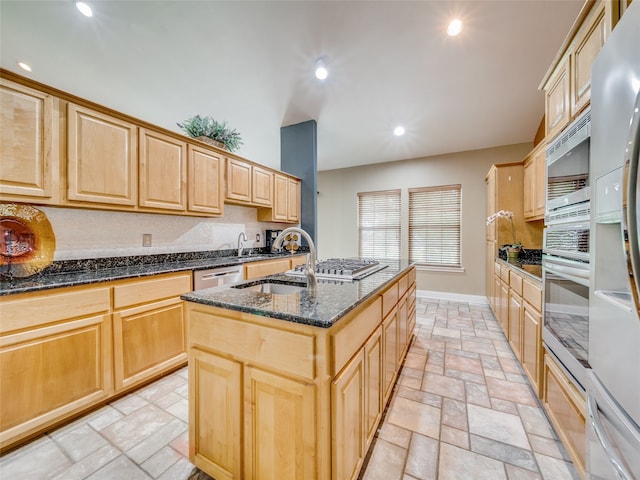 kitchen featuring stainless steel appliances, sink, light brown cabinets, dark stone countertops, and a center island with sink