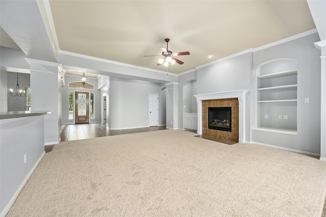 unfurnished living room featuring built in shelves, ceiling fan with notable chandelier, decorative columns, light colored carpet, and a tiled fireplace