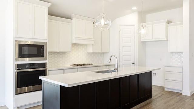 kitchen with an island with sink, sink, pendant lighting, light wood-type flooring, and appliances with stainless steel finishes