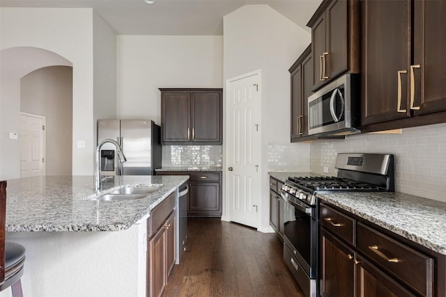 kitchen featuring appliances with stainless steel finishes, light stone countertops, an island with sink, tasteful backsplash, and dark hardwood / wood-style flooring