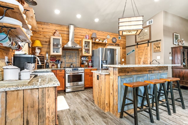 kitchen with a barn door, a kitchen island with sink, stainless steel appliances, wall chimney exhaust hood, and glass insert cabinets