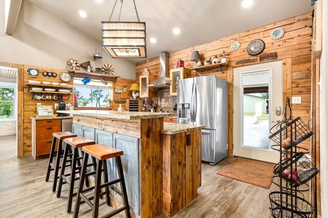 kitchen featuring wood walls, light hardwood / wood-style flooring, stainless steel refrigerator with ice dispenser, and wall chimney exhaust hood