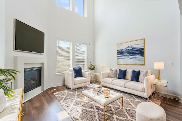 living area with dark wood-type flooring, a glass covered fireplace, a high ceiling, and baseboards