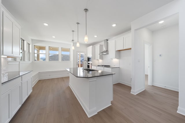 kitchen featuring white cabinetry, wall chimney exhaust hood, an island with sink, pendant lighting, and light hardwood / wood-style floors