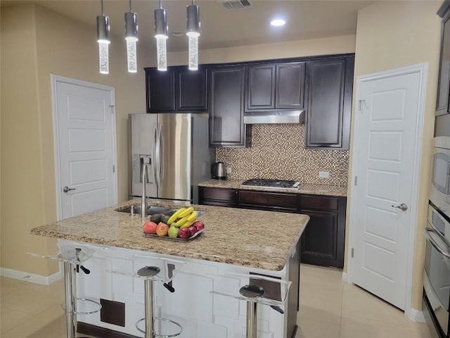 kitchen featuring appliances with stainless steel finishes, a kitchen island with sink, light stone counters, tasteful backsplash, and decorative light fixtures