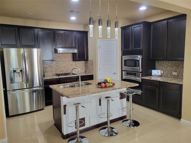 kitchen with sink, appliances with stainless steel finishes, hanging light fixtures, light stone counters, and a center island with sink