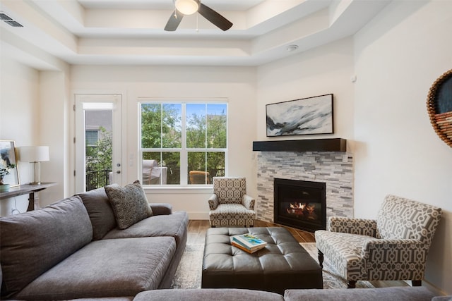 living room featuring hardwood / wood-style floors, a stone fireplace, a raised ceiling, and ceiling fan