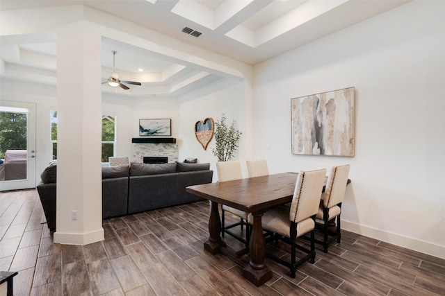 dining space with a tray ceiling, a stone fireplace, ceiling fan, and dark wood-type flooring