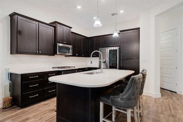 kitchen featuring appliances with stainless steel finishes, light wood-type flooring, sink, a center island with sink, and hanging light fixtures