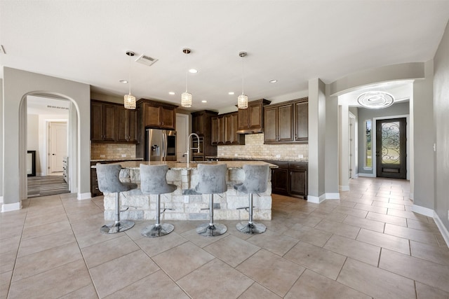 kitchen featuring backsplash, stainless steel fridge with ice dispenser, a kitchen island with sink, light stone countertops, and decorative light fixtures