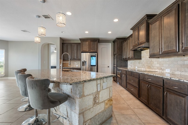 kitchen featuring stainless steel fridge, a kitchen island with sink, dark brown cabinetry, and pendant lighting