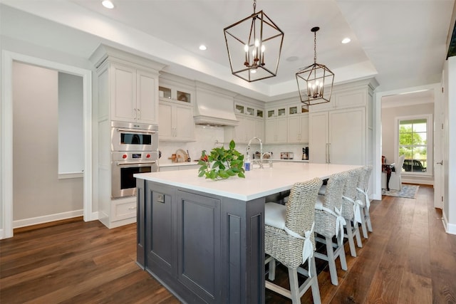 kitchen with a center island with sink, dark hardwood / wood-style floors, custom exhaust hood, a tray ceiling, and double oven