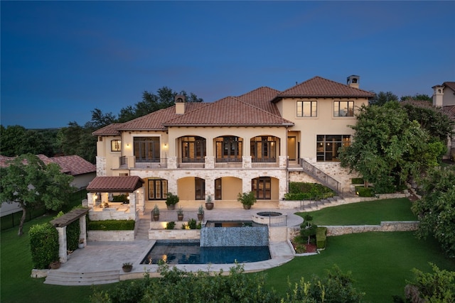 back house at dusk featuring a yard, a pool with hot tub, a balcony, and a patio area