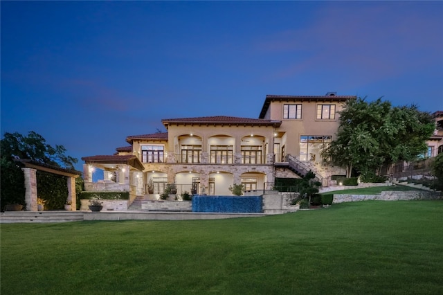 back house at dusk with a balcony, a lawn, and a patio