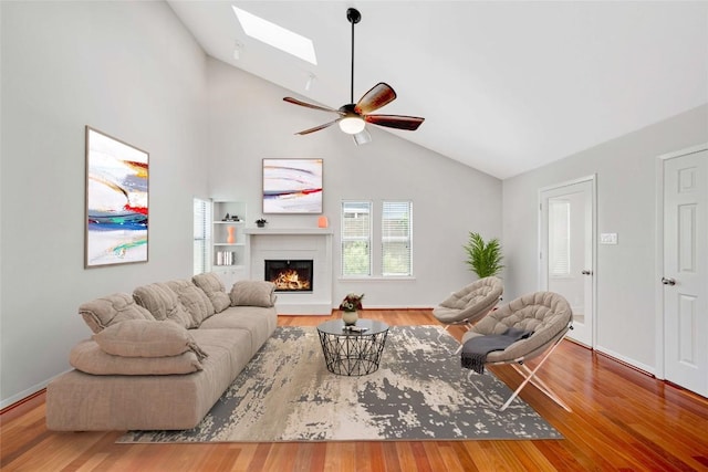 living area featuring vaulted ceiling with skylight, baseboards, ceiling fan, wood finished floors, and a lit fireplace